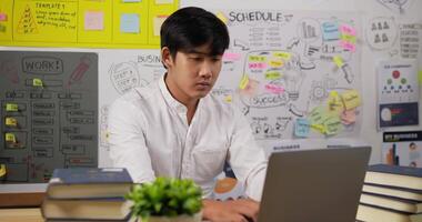 porträtt av asiatisk affärsman att skriva laptop medan han sitter vid skrivbordet på arbetsplatsen på hemmakontoret. trött manlig arbetare gäspar arbetar på anteckningsboken. uttråkad anställd som arbetar på kontor koncept. video