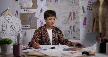 Asian woman fashion designer contemplating drawings and working on laptop in the studio. Female clothing designer sitting at a desk in her office. SME marketing and entrepreneur concept.