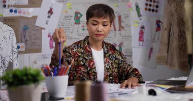 retrato de designer de moda de mulher asiática desenha um esboço de roupas femininas enquanto está sentado no estúdio. feliz empresária de inicialização está em processo de criação de uma nova coleção de roupas. video