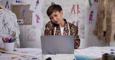 porträtt av asiatisk kvinna modedesigner pratar i mobiltelefon med kunden medan du arbetar på laptop i studion. glad nystartad småföretagare. sme marknadsföring och entreprenörskoncept. video