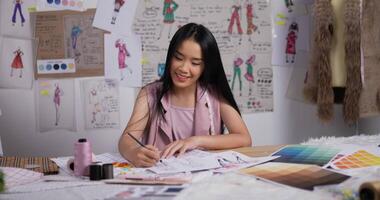 el retrato de una diseñadora de moda asiática dibuja un boceto de ropa de mujer mientras se sienta en el estudio. feliz empresaria nueva está en proceso de crear una nueva colección de ropa. video