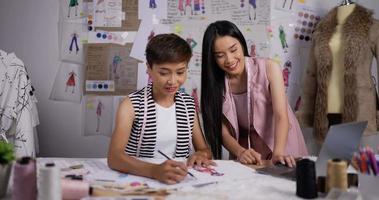 porträtt av glada två asiatiska modedesigner kvinnor som arbetar i studio. unga flickor kollegor skissa och titta på laptop. video