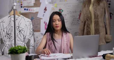 retrato de designer de moda de mulher asiática falando no celular com o cliente enquanto trabalhava no laptop no estúdio. proprietário de uma pequena empresa de inicialização ocupada. sme marketing e conceito de empresário.