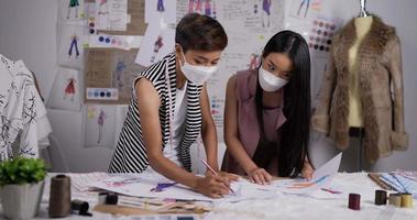 Porträt von zwei asiatischen Modedesignerinnen, die eine medizinische Gesichtsmaske tragen, zeichnet eine Skizze von Damenbekleidung im Studio. Eine glückliche Startup-Geschäftsfrau ist dabei, eine neue Kleiderkollektion zu erstellen. video