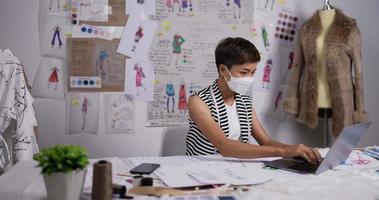 retrato da mulher estilista asiática usa máscara facial médica trabalhando no laptop e falando ao telefone com o cliente no estúdio. conceito de negócio sme. video