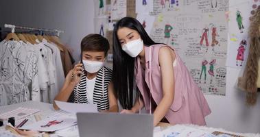 retrato de duas mulheres asiáticas designer de moda usam máscara facial médica olhando um esboço de roupas femininas no laptop e falando ao telefone com o cliente no estúdio. conceito de negócio sme. video