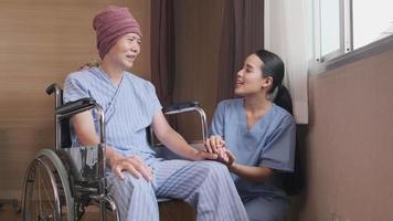 Uniformed young Asian female therapy doctor holding hands wheelchair male patient at window to support and motivate recovery, cancer illness after chemo medical treatment in hospital inpatient room.