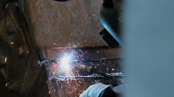 A man wearing a welding mask and gloves works in a home workshop with a welding machine. Worker welding metal with sparks, close-up video