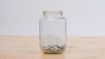 Stop motion animation coin into a clear glass jar on wooden desk. Savings and Investments concept. video