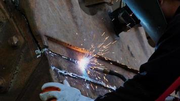 A man wearing a welding mask and gloves works in a home workshop with a welding machine. Worker welding metal with sparks, close-up video