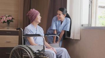 Uniformed young Asian female therapy doctor encouraging wheelchair male patient at window to support and motivate recovery, cancer illness after chemo medical treatment in hospital inpatient room. video