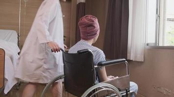 Una joven y uniformada doctora de terapia caucásica empuja a un paciente varón en silla de ruedas a la ventana, apoya y motiva la recuperación de la enfermedad del cáncer después del tratamiento médico con quimioterapia en la habitación del hospital. video