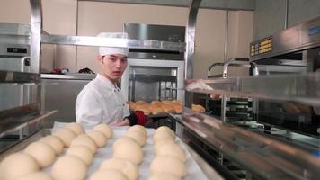 joven chef asiático profesional con uniforme de cocinero blanco con sombrero, guantes y delantal haciendo pan con masa de pastelería, preparando comida fresca de panadería, horneando en el horno en la cocina de acero inoxidable del restaurante. video