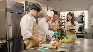 curso de cocina de hobby, chef masculino senior en uniforme de cocinero enseña a los jóvenes estudiantes de cocina a preparar, mezclar y revolver ingredientes para pasteles, pasteles de frutas en la cocina de acero inoxidable del restaurante. video