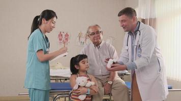 un médico caucásico profesional asesora al abuelo y explica los medicamentos recetados a los médicos en formación en una cama de hospital para pacientes, consultas de enfermedades y clínica de examen de salud familiar. video