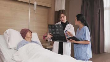 une femme médecin caucasienne en diagnostic uniforme explique le film radiographique avec un radiologue asiatique et un patient de sexe masculin en convalescence au lit d'une chambre d'hôpital dans une salle d'hôpital, une clinique médicale, une consultation d'examen du cancer. video