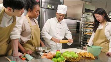 Cuisine course, senior male chef in cook uniform teaches young cooking class students to knead pastry dough and prepare ingredients for bakery foods, fruit pies in restaurant stainless steel kitchen. video
