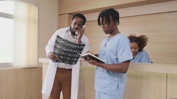 Professional African-American male doctor talks phone about a patient's diagnosis X-ray film and medicine at reception counter of hospital's outpatient clinic, and young medical students together. video