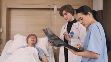 Caucasian female doctor in uniform diagnosis explains x-ray film with Asian radiologist and recovery male patient at inpatient room bed in a hospital ward, medical clinic, cancer examination consult. video