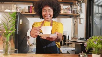 African American female barista in looks at camera, offers cup of coffee to customer with cheerful smile, happy service works in casual restaurant cafe, young small business startup entrepreneur.