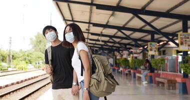 seitenansicht eines jungen asiatischen reisepaares, das am bahnhof auf den zug wartet. mann und frau mit schutzmasken während des notfalls covid-19. transport-, reise- und soziales distanzierungskonzept. video