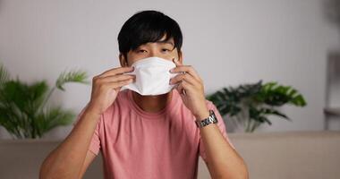 Portrait of Smiling Asian man wearing face mask sits on couch in living room. Happy 20s man putting on protective face masks for covid-19. Coronavirus protection concept. video