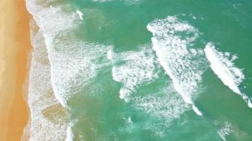 vista aérea de la playa de arena y la textura de la superficie del agua. olas espumosas con cielo. vuelo de drones de una hermosa playa tropical. increíble costa de arena con olas de mar blanco. concepto de naturaleza, paisaje marino y verano. video