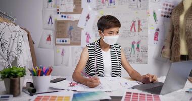 portret van aziatische modeontwerper vrouw draagt een masker tekent een schets van dameskleding en werkt op laptop in de studio. zakenvrouw is bezig met het maken van een nieuwe kledingcollectie. video