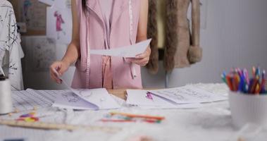 Porträt eines jungen asiatischen Modedesigners oder Stylisten, der im Studio Skizzen für das neue Kleid auswählt. schneiderinnen bereiten garderobenkleidung zum verkauf vor. video