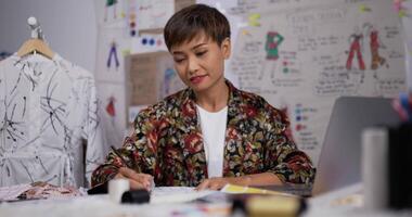 Portrait of Asian woman fashion designer draws a sketch of women's clothing while sitting at the studio. Happy startup small businesswoman is in process of creating a new clothes collection. video