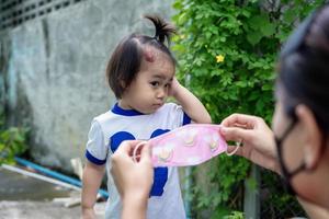 Adorable little child girl with big capillary strawberry hemangiomas red birthmark on head refuses to wear medical face mask that the mother or guardian are trying to put on face. Health care concept photo