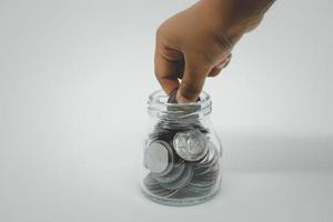 mano de niño poniendo o recogiendo monedas de baht de un frasco de vidrio con fondo blanco. concepto de ahorro de dinero. foto