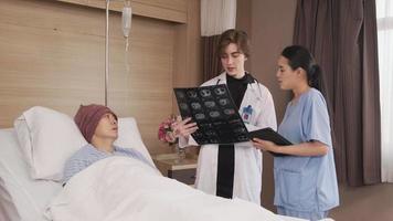 une femme médecin caucasienne en diagnostic uniforme explique le film radiographique avec un radiologue asiatique et un patient de sexe masculin en convalescence au lit d'une chambre d'hôpital dans une salle d'hôpital, une clinique médicale, une consultation d'examen du cancer. video