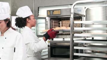 Professional African American female chef in white cook uniform, gloves, and apron making bread from pastry dough, preparing fresh bakery food, baking in oven at restaurant's stainless steel kitchen. video