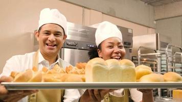 portrait de chefs professionnels en uniforme blanc regardant la caméra avec un sourire joyeux et fier avec un plateau de pain dans la cuisine. un ami et partenaire des aliments de boulangerie et de l'occupation quotidienne de la boulangerie fraîche. video