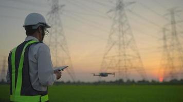 An electrical engineer is using a remote control drone to inspect high-voltage poles during sunset or rise.