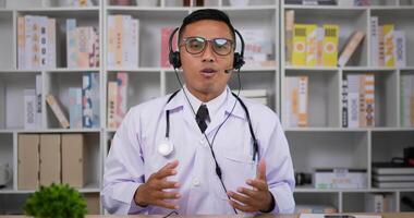 portrait d'un médecin de sexe masculin asiatique professionnel en blouse médicale blanche. casque d'homme faisant une conférence téléphonique et regardant la caméra. consultation patient à distance en ligne par webcam. notion de télémédecine. video