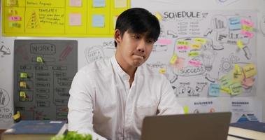 retrato do empresário asiático pensativo pensando e trabalhando no laptop enquanto está sentado na mesa do local de trabalho no escritório em casa. homem freelancer trabalhando em casa. navegando na internet, usando redes sociais. video