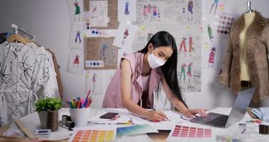 retrato da máscara de desgaste da mulher de designer de moda asiática desenha um esboço de roupas femininas e trabalhando no laptop no estúdio. empresária está em processo de criação de uma nova coleção de roupas.