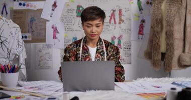 asiatische modedesignerin, die zeichnungen erwägt und im studio am laptop arbeitet. weibliche kleidungsdesignerin, die an einem schreibtisch in ihrem büro sitzt. KMU-Marketing und Unternehmerkonzept. video