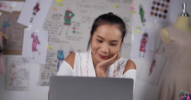 créatrice de mode femme asiatique envisageant des dessins via un ordinateur portable dans le studio. heureuse créatrice de vêtements féminine travaillant assise à un bureau dans son bureau. marketing des PME et concept d'entrepreneur. video