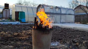 Garbage burns in a metal tank on the background of a greenhouse
