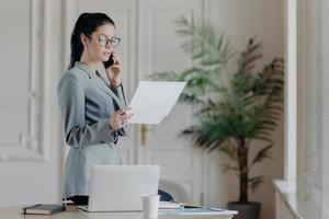 Serious brunette female employee stands near desktop, holds paper documents, talks via smartphone, wears formal clothes and spectacles, discusses business strategy with partner, organizes meeting photo