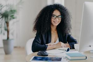 Photo of successful female manager in elegant suit, smiles pleasantly, makes notes, sits at desktop, drinks coffee, poses at computer, smiles positively, poses in office interior. People and work