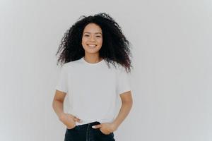 Isolated shot of young African American woman wears white t shirt, expresses good emotions, stands alone indoor, poses for photo, has casual talk with friend, enjoys free time. People and happiness photo