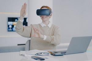 Girl is sitting at the desk in 3d goggles in front of laptop. Concept of modern devices using. photo