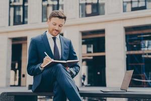 Smiling young man dressed in blue suit taking notes in notebook while spending time outdoors photo