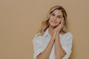 Romantic young blonde lady in casual white shirt keeping folded hands near her face photo