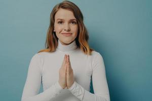 joven suplicó mujer confiada posando con las manos dobladas en gesto de oración, aislada en azul foto