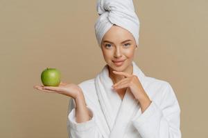Beautiful healthy woman touches chin gently holds green apple has perfect smooth skin wears soft white domestic robe and wrapped towel on head models indoor with fresh fruit. Skin treatment. photo